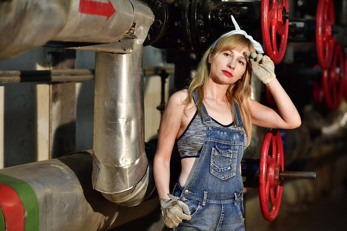 Femme en overalls et brassière devant tuyaux industriels et roues rouges, pose réflexive
